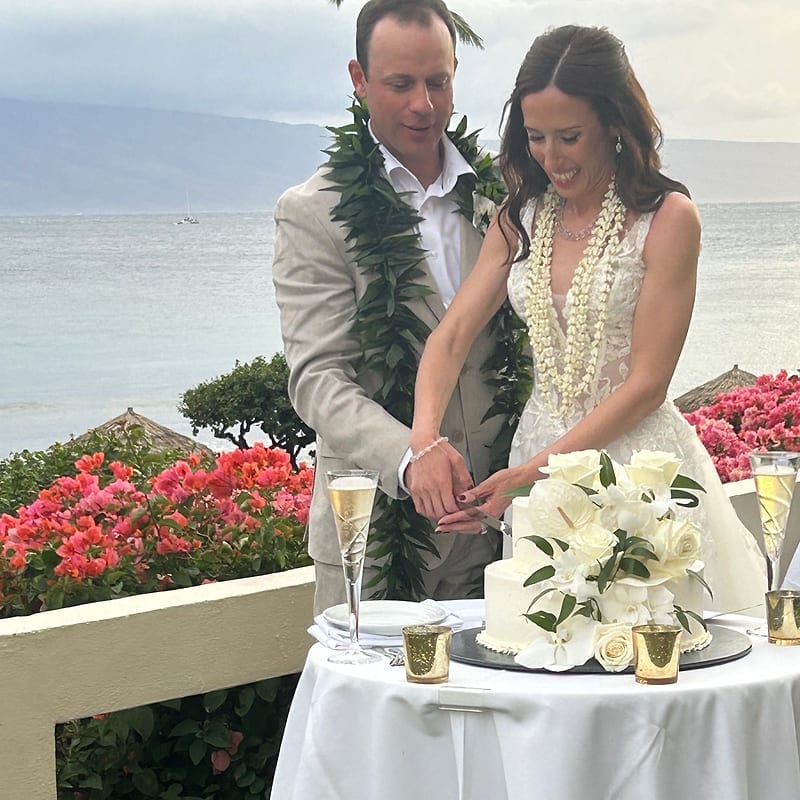 Bride and groom in leis cutting a white floral wedding cake at a seaside reception