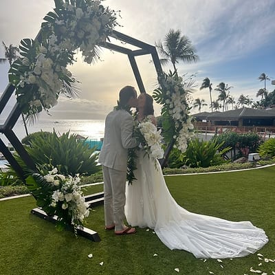 Bride and groom kissing under a tropical floral arch on a lawn overlooking the ocean